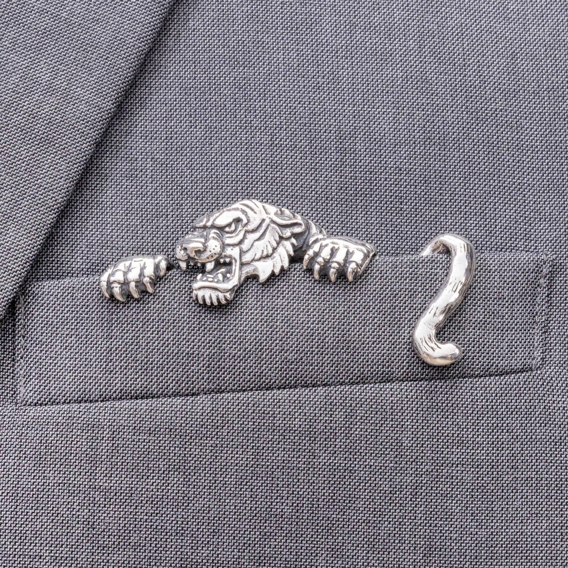 A sterling silver pocket square with a carved tiger design inserted into a jacket pocket, displayed on a gray fabric surface.