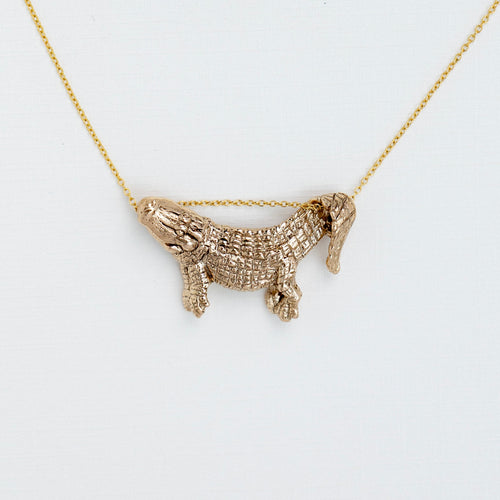 A gold-toned necklace featuring a textured alligator pendant on a delicate chain, photographed against a clean white background.