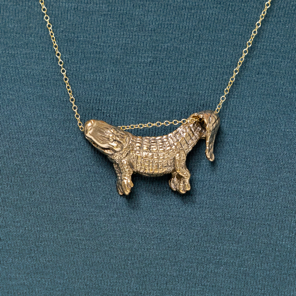 A bronze dangling alligator pendant on a chain, displayed against a blue background.