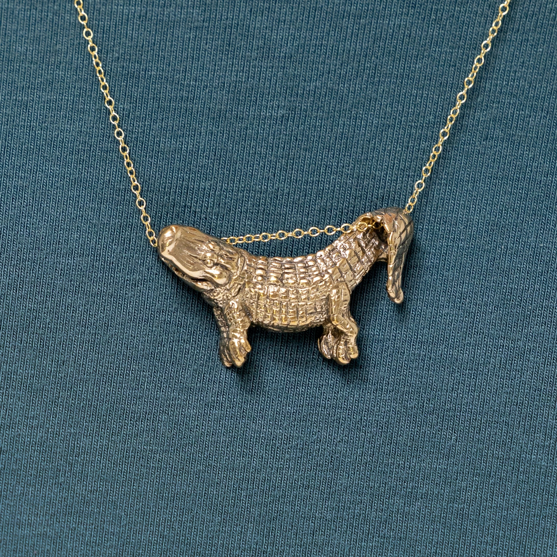 A bronze dangling alligator pendant on a chain, displayed against a blue background.
