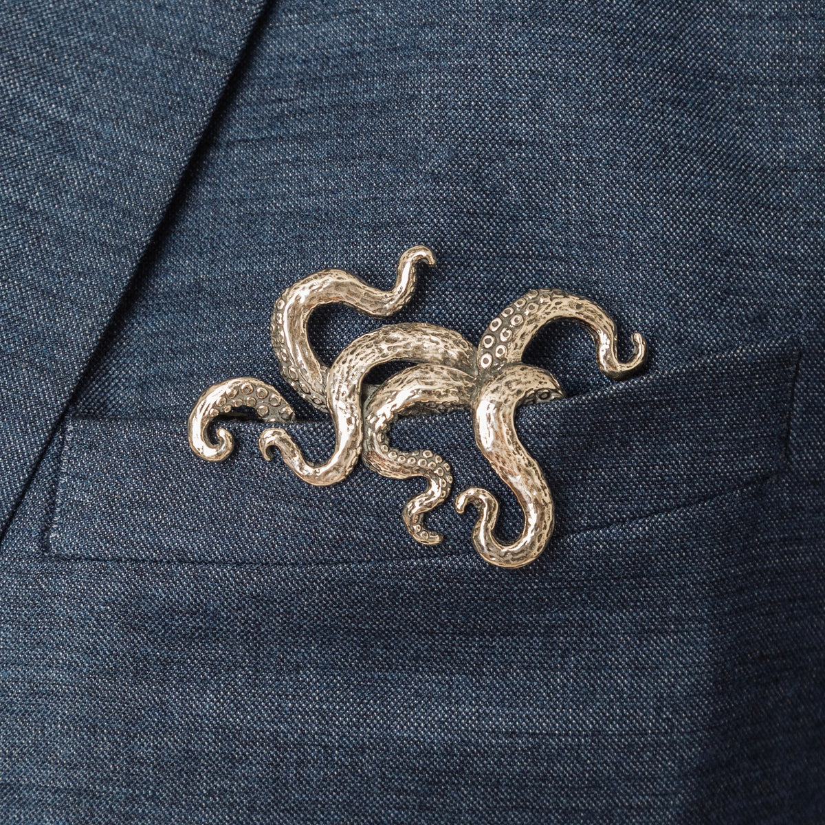 A silver-colored, hand-carved octopus (Kraken) pocket square insert protruding from a suit jacket pocket.